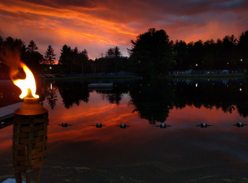 A serene lake at sunset, reflecting vibrant orange and purple skies, with a lit tiki torch in the foreground.