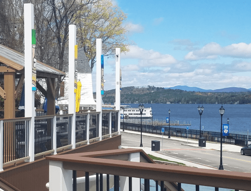 View of a lakeside promenade with colorful poles, a boat on the water, and mountains in the background under a blue sky.
