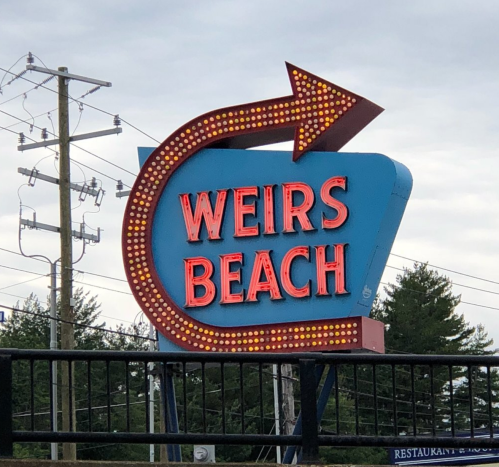 Brightly lit sign for Weirs Beach with an arrow, set against a cloudy sky and power lines in the background.