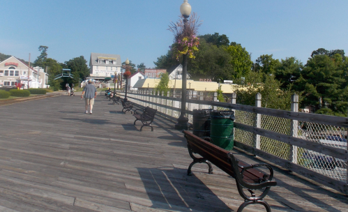 A wooden boardwalk lined with benches, trees, and buildings, with a person walking in the distance on a sunny day.