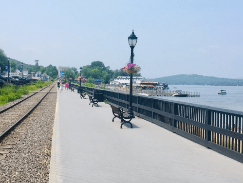 A scenic waterfront walkway with benches, flowers, and boats, alongside a railway track on a sunny day.