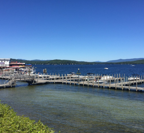 A sunny day at a lakeside dock with boats and a clear blue sky, surrounded by green hills in the background.