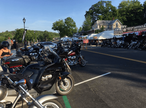 A row of parked motorcycles along a street with vendors and trees in the background on a sunny day.