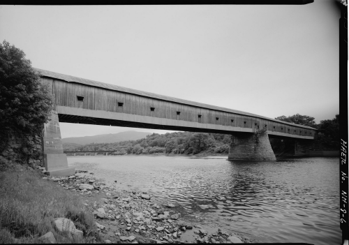 A historic covered bridge spans a river, surrounded by lush greenery and a cloudy sky. Black and white photo.
