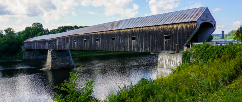 A long wooden covered bridge spans a calm river, surrounded by greenery and under a blue sky with scattered clouds.
