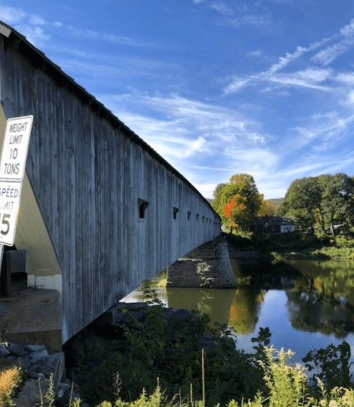 A wooden covered bridge spans a calm river, with a weight limit sign and trees in the background under a blue sky.