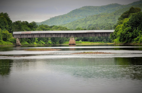 A wooden covered bridge spans a calm river, surrounded by lush green hills and trees under a cloudy sky.