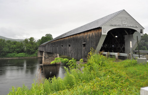 A wooden covered bridge spans a calm river, surrounded by lush greenery and overcast skies.