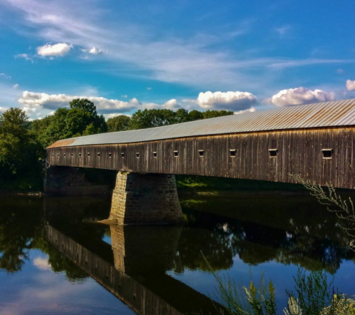 A wooden covered bridge spans a calm river, reflecting the blue sky and clouds above. Lush greenery surrounds the scene.