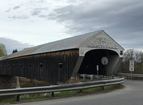 A weathered covered bridge with a metal roof spans a river, surrounded by trees under a cloudy sky.