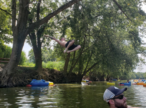 A person jumps off a tree into a river, while others float on colorful tubes nearby.