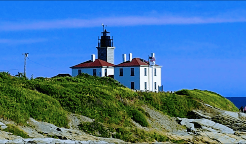 A white lighthouse with a red roof stands on a grassy hill by the ocean under a clear blue sky.