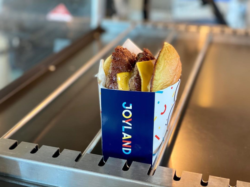 A colorful box labeled "JOYLAND" holds a delicious-looking hot dog topped with cheese and crispy fried chicken.