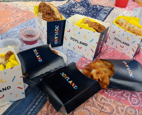 Five takeout boxes from Joyland filled with fried chicken and sides, placed on a colorful patterned tablecloth.