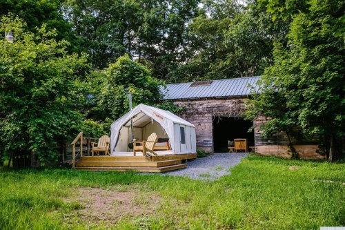 A cozy glamping tent on a wooden deck, surrounded by lush greenery and an old barn in the background.