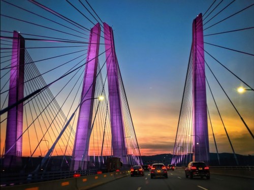A vibrant sunset behind a cable-stayed bridge, illuminated by purple lights, with cars driving across.