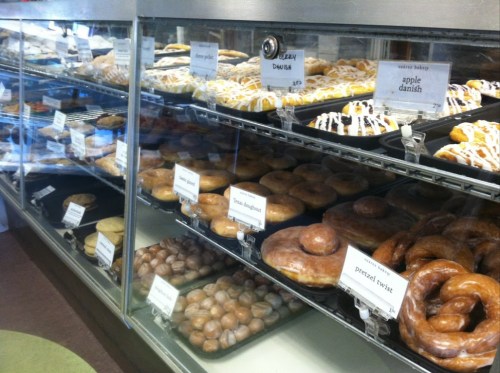 A display case filled with various pastries, including donuts, danishes, and pretzel twists, all labeled with prices.