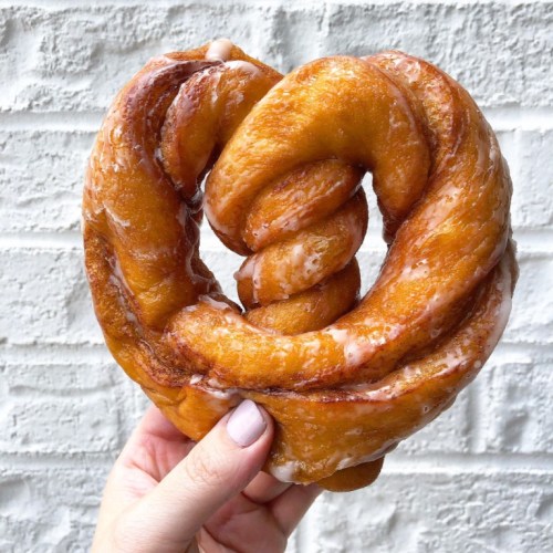 A hand holding a large, heart-shaped glazed pretzel against a white brick background.