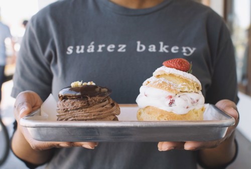 A person holds a tray with two desserts: a chocolate mousse and a strawberry shortcake, wearing a "Suárez Bakery" shirt.