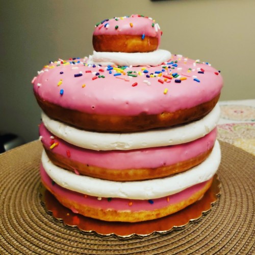 A colorful, layered cake resembling donuts, topped with pink icing and sprinkles, with a smaller donut on top.
