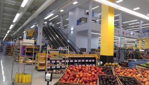 A grocery store interior featuring colorful fruit displays, shelves of products, and escalators in the background.
