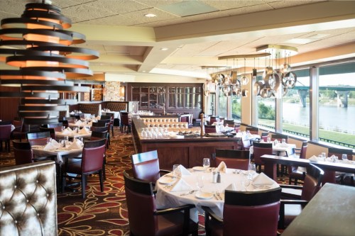 Elegant restaurant interior with tables set for dining, large windows overlooking a river, and modern light fixtures.