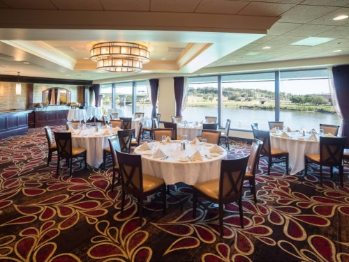 Elegant dining room with round tables set for a meal, large windows overlooking a river, and decorative carpet.