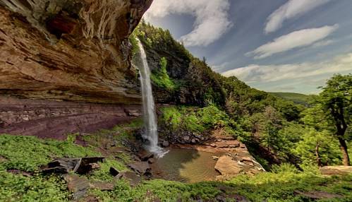 A scenic waterfall cascades down rocky cliffs into a serene pool, surrounded by lush greenery and a blue sky.