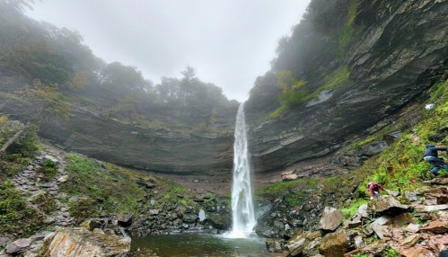 A tall waterfall cascades down rocky cliffs, surrounded by lush greenery and misty fog.