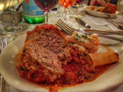 A plate of meatballs in marinara sauce with rigatoni pasta, garnished with herbs and cheese, alongside drinks.