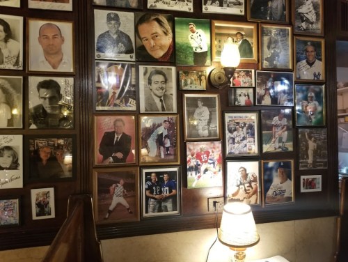 A wall covered with framed photos of various people, including athletes and celebrities, illuminated by a lamp.