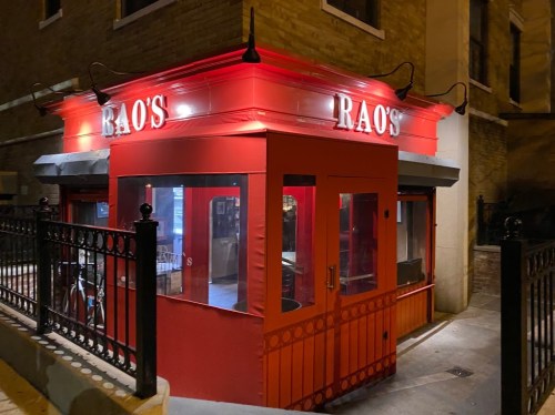 A red restaurant exterior with the name "Rao's" illuminated at night, featuring large windows and a decorative entrance.