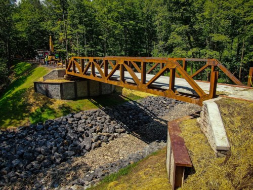 A wooden bridge spans a rocky area, surrounded by lush green trees and a construction site in the background.