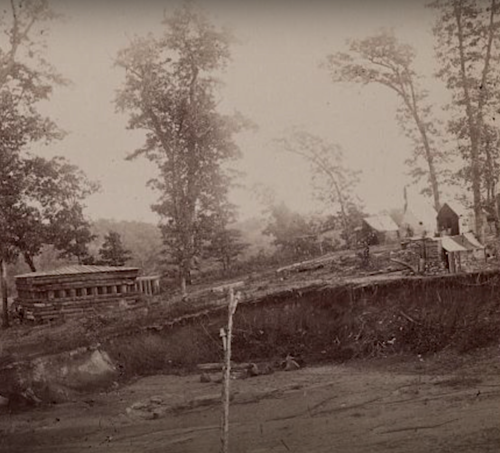 Historic black-and-white photo of a rural settlement with wooden structures and tents among trees.