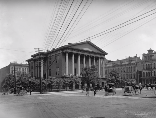 Historic building with columns, surrounded by trees and horse-drawn carriages, set in a bustling urban scene.