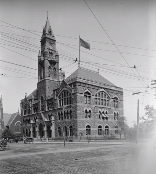 Historic brick building with a tall clock tower and flag, surrounded by power lines and trees. Black and white photo.