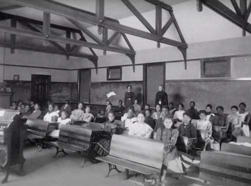 Historic black-and-white photo of a classroom with students and teachers seated at wooden desks.
