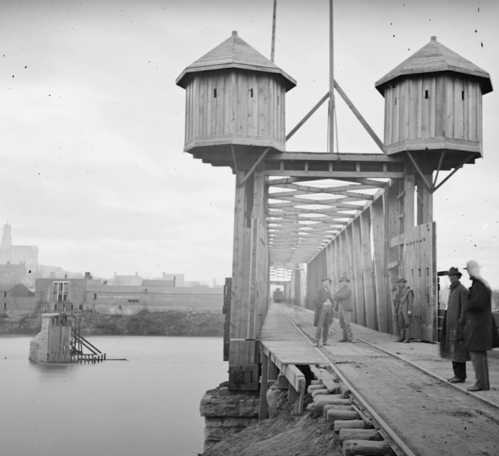 Historic black-and-white photo of a wooden bridge under construction, featuring two towers and workers on the structure.