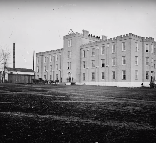 Historic building with a castle-like design, surrounded by a grassy area and a few horses nearby. Black and white photo.