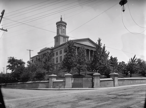 Historic building with a tall tower, surrounded by trees and a stone wall, captured in black and white.