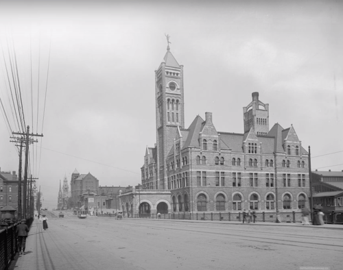 Historic black-and-white photo of a grand brick building with a clock tower, set on a snowy street.