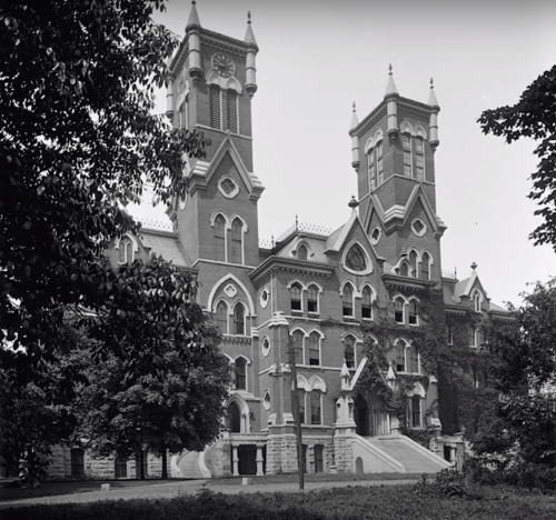 Historic brick building with towers, intricate architecture, and surrounding trees, captured in black and white.