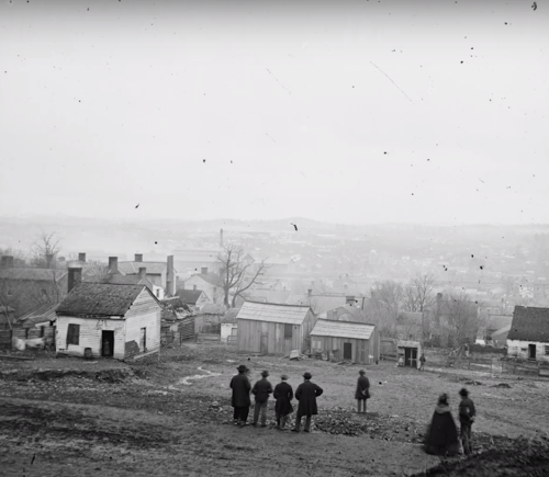 Black and white photo of a rural landscape with several people standing near simple houses, overlooking a valley.