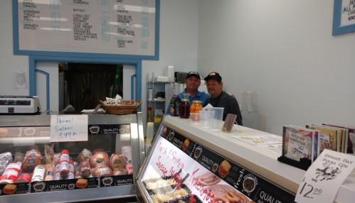 Two men stand behind a deli counter filled with various meats and products, smiling at the camera.