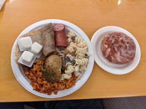 A plate of food with rice, pasta, sausage, bread, and a donut on the side, all served on disposable plates.