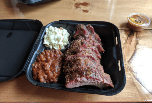 A takeout container with sliced smoked brisket, coleslaw, baked beans, and a small cup of sauce on a wooden table.