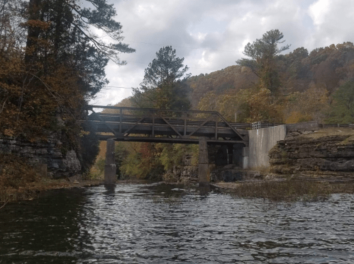 A wooden bridge spans a river, surrounded by rocky banks and autumn-colored trees under a cloudy sky.