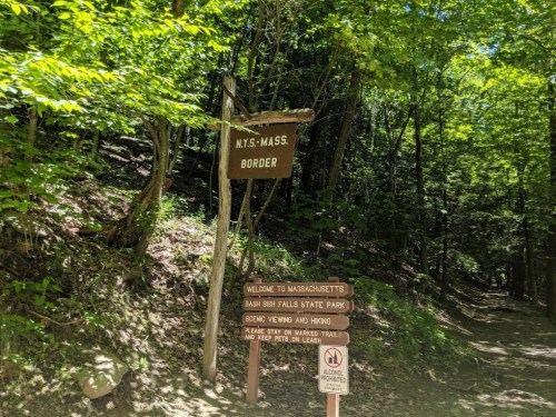Sign marking the New York-Massachusetts border near Bash Bish Falls State Park, surrounded by lush greenery.