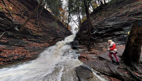 A waterfall cascades down rocky cliffs, surrounded by trees with autumn foliage, while a person stands nearby.