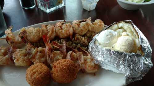 A plate of grilled shrimp, rice, a baked potato with butter, and two fried balls.